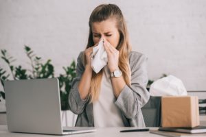 blonde businesswoman sneezing in tissue near laptop in office